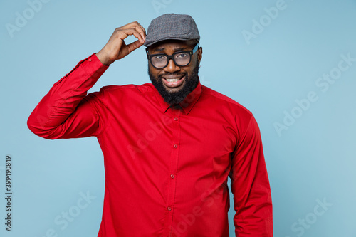 Preoccupied puzzled young bearded african american man 20s wearing casual red shirt eyeglasses cap standing put hand on head looking camera isolated on pastel blue color background studio portrait. © ViDi Studio