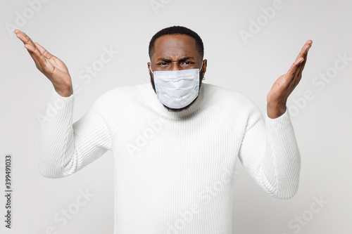 Perplexed young african american man in basic sweater sterile face mask to safe from coronavirus virus covid-19 during pandemic quarantine spreading hands isolated on white background studio portrait. photo