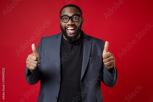 Valokuva Excited cheerful young african american business man 20s wearing classic jacket suit eyeglasses standing showing thumbs up looking camera isolated on bright red color wall background studio portrait