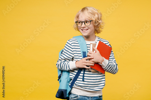 Smiling funny little male teen boy 10s years old in striped sweatshirt eyeglasses backpack hold school books looking aside isolated on yellow color background child studio portrait. Education concept.