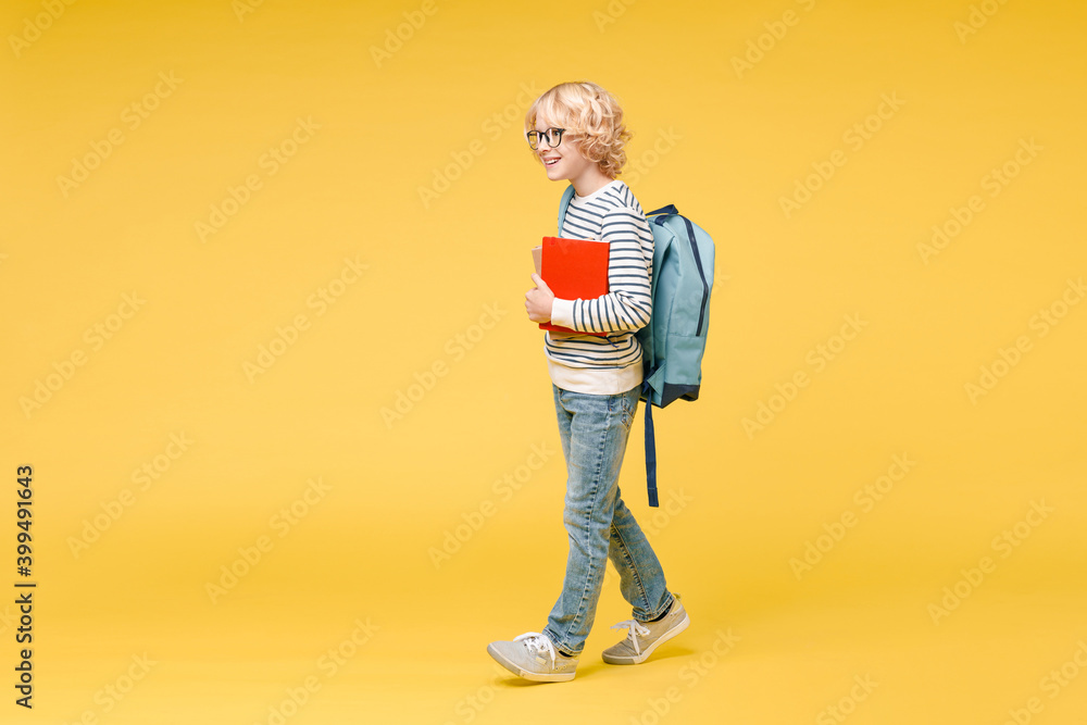Fototapeta premium Full length side view of pretty male kid teen boy 10s in striped sweatshirt eyeglasses backpack hold school books walking going isolated on yellow background child studio portrait. Education concept.