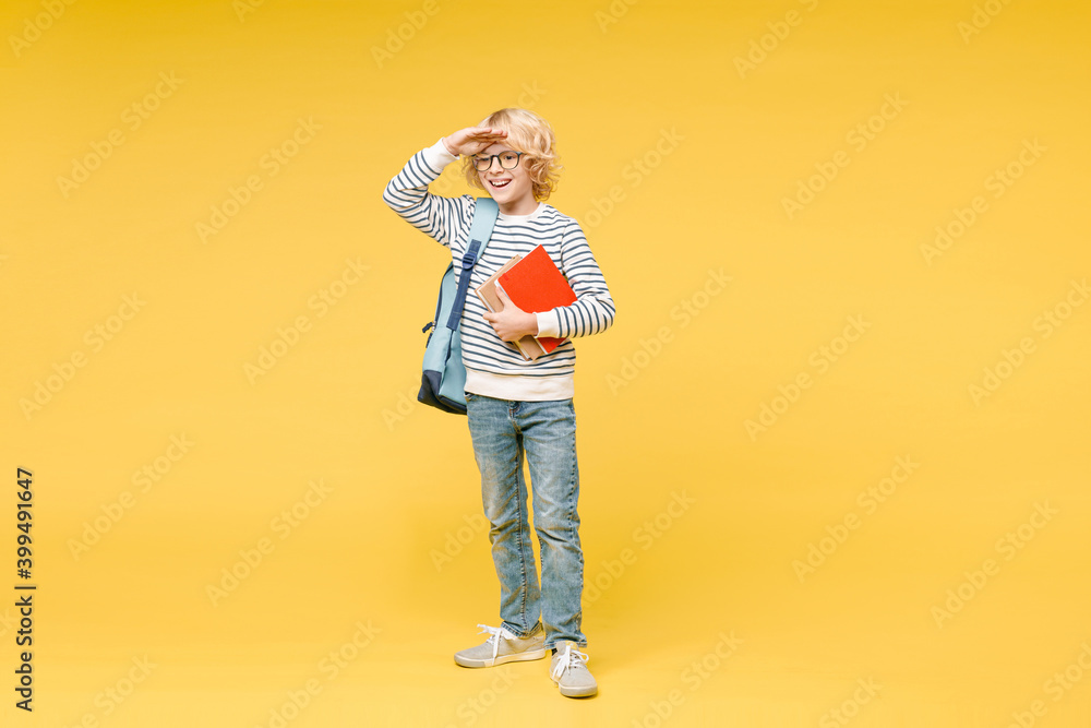 Fototapeta premium Full length of cheerful male boy 10s in sweatshirt eyeglasses backpack hold school books put hand at forehead looking far away distance isolated on yellow background child portrait. Education concept.