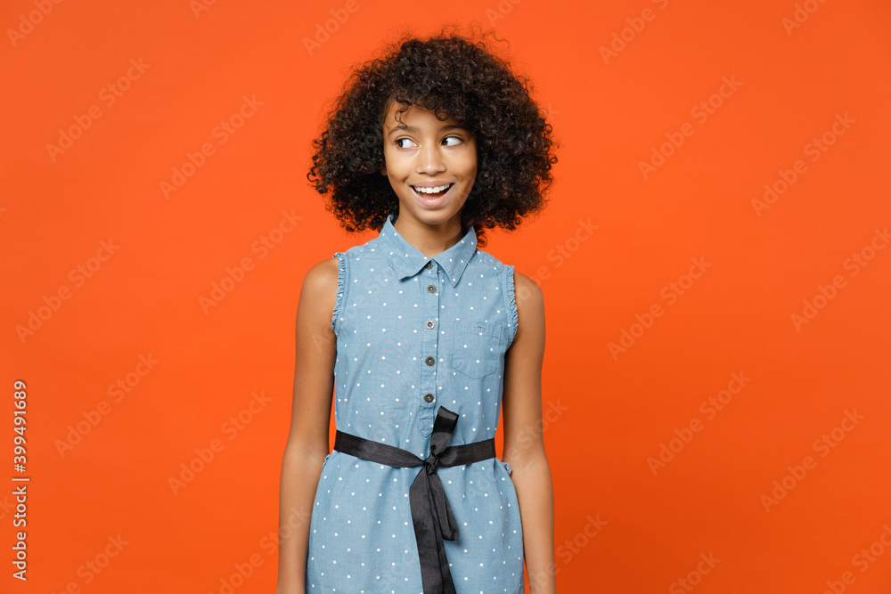 Excited little african american kid girl 12-13 years old wearing casual denim dress looking aside isolated on bright orange color background children studio portrait. Childhood lifestyle concept. Excited little african american kid girl 12-13 years old wearing casual denim dress looking aside isolated on bright orange color background children studio portrait. Childhood lifestyle concept.