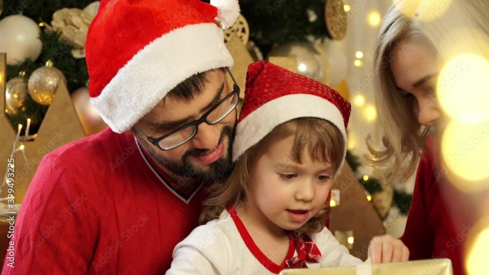 Portrait of happy young family celebrate Christmas and New year. Smiling people, father and mother in red santa hats give gift to child girl on background of Christmas tree. Close up Portrait of happy young family celebrate Christmas and New year. Smiling people, father and mother in red santa hats give gift to child girl on background of Christmas tree. Close up