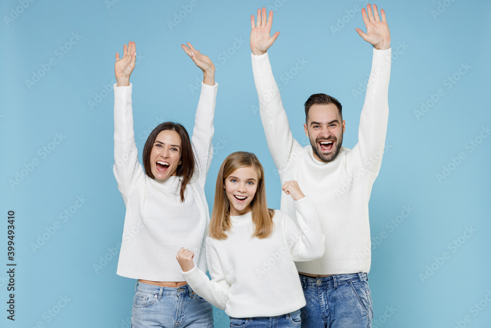 Obraz premium Happy excited young parents mom dad with child kid daughter teen girl in white sweaters doing winner gesture rising hands isolated on blue background studio portrait. Family day parenthood concept.