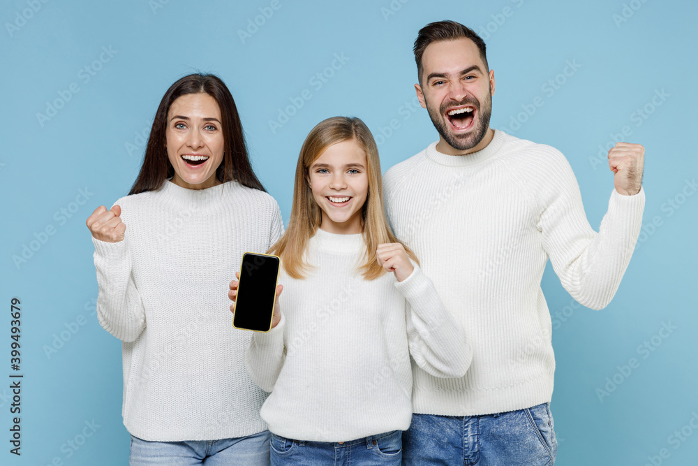 Happy young parents mom dad with child kid daughter teen girl in sweaters doing winner gesture hold mobile phone with blank empty screen isolated on blue background. Family day parenthood concept.