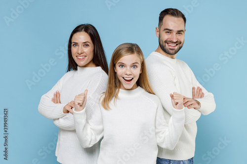 Excited young parents mom dad with child kid daughter teen girl in white sweaters hold hands crossed doing winner gesture isolated on blue background studio. Family day parenthood childhood concept. photo