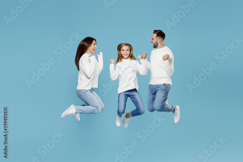 Full length of joyful young parents mom dad with child kid daughter teen girl in white sweaters jumping doing winner gesture isolated on blue background studio portrait. Family day parenthood concept.