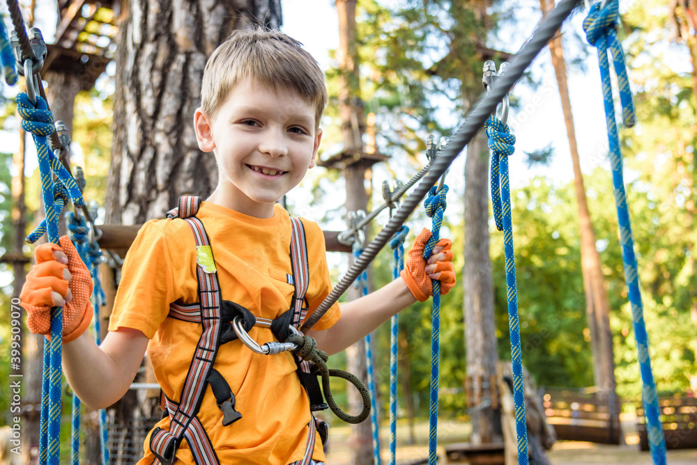 Young cute child boy in summer clothing, safety harness and helmet attached with carbine to cable moves slowly along rope way on green trees sunny bokeh background. Sport, game, leisure concept Young cute child boy in summer clothing, safety harness and helmet attached with carbine to cable moves slowly along rope way on green trees sunny bokeh background. Sport, game, leisure concept