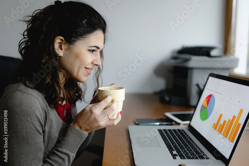 Close-up young caucasian business woman portrait working at office desk. Smiling lady holding a cup of hot drink while studying graphs on desktop computer.