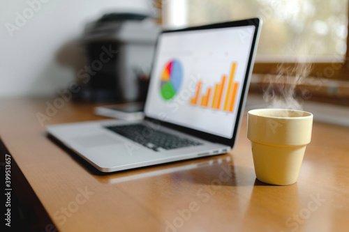 Close-up workplace with computer laptop and cup of hot steaming drink. Histograms on pc screen. Growth, analysis, comfortable casual tele working concept.