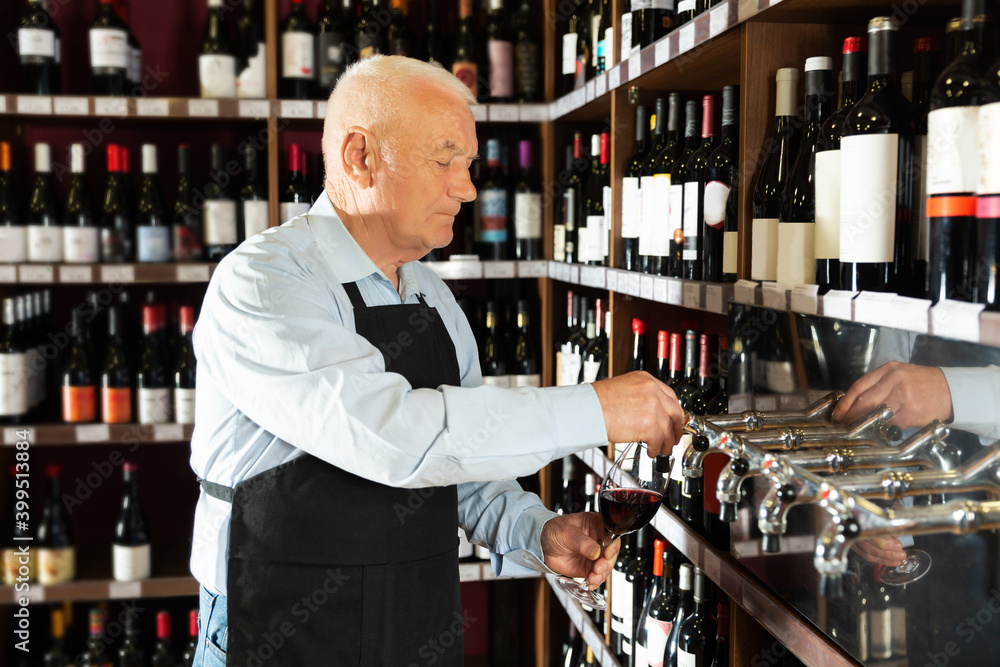 Portrait of positive senior man professional sommelier pouring wine from wine column