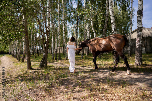 A woman in a white dress and barefoot leads a horse by the bridle. Back view. © Aleksandr