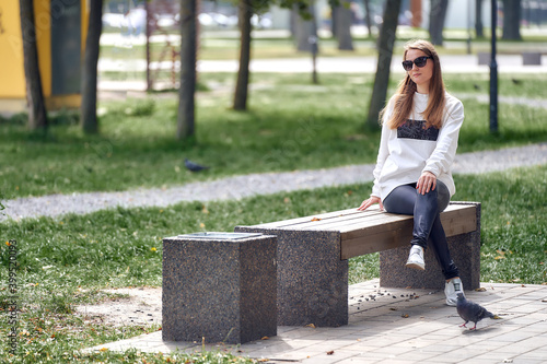 Young woman in sunglasses sits on a wooden bench in the park.