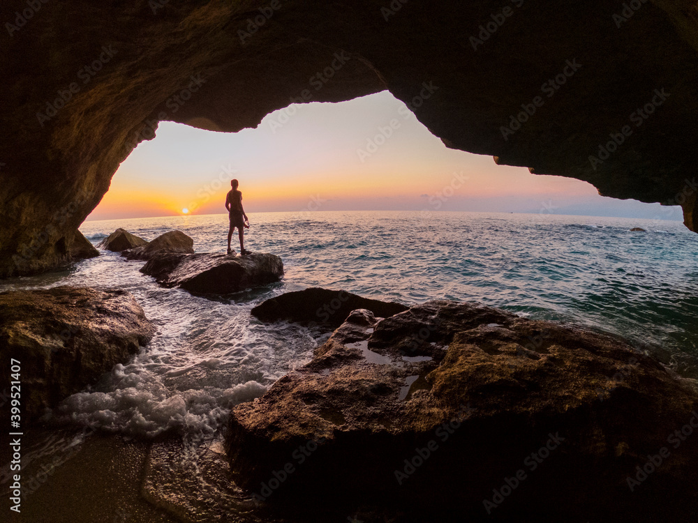 
Man from behind against the light inside a cave observes the sunset overlooking the sea, warm and enveloping colors, image of strong emotional impact