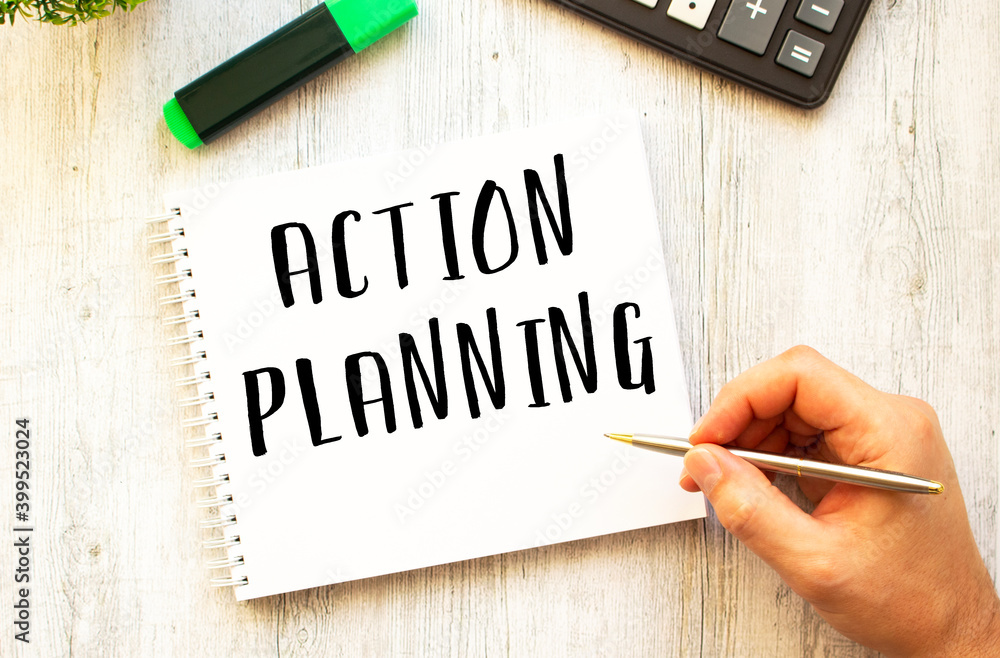 A hand with a pen writes text ACTION PLANNING in a notebook on a wooden desk. View from above. A hand with a pen writes text ACTION PLANNING in a notebook on a wooden desk. View from above.