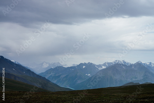 Gloomy view to great snowy mountains under dark gray cloudy sky. Dramatic alpine landscape with snow mountains in rainy weather. Atmospheric scenery with giant mountain ridge in overcast darkness.