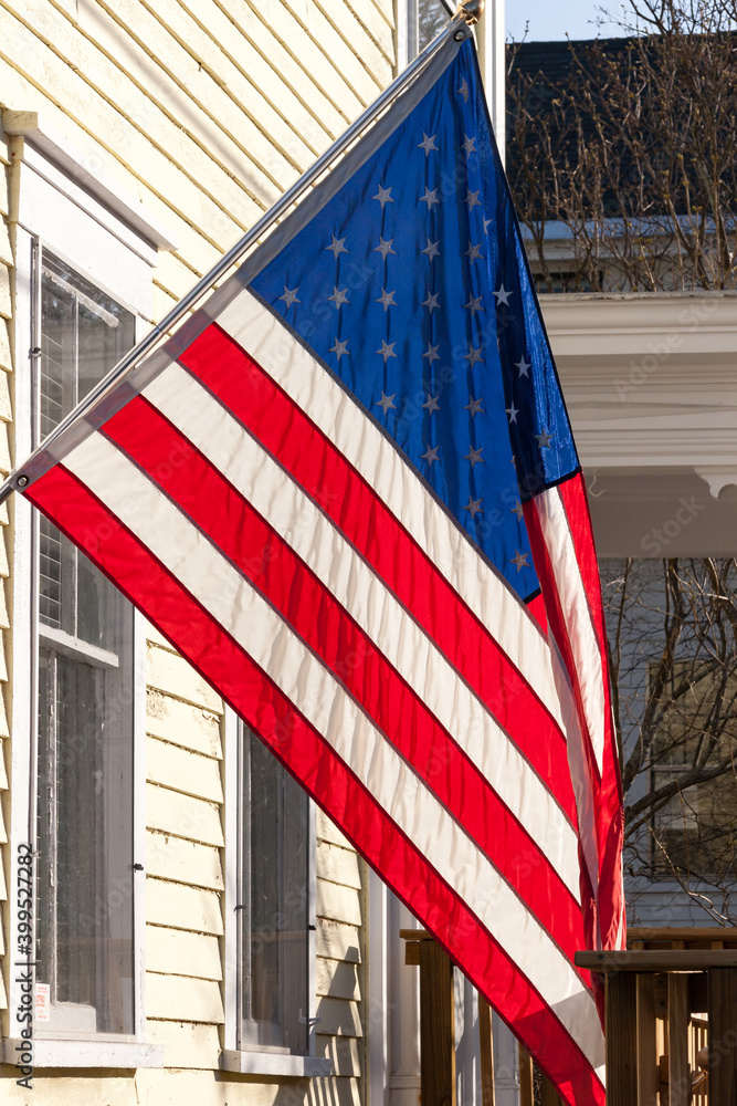 A close up view of an American stars and stripes flag hanging diagonally in front of a white wooden home displaying pride and patriotism. New Hampshire, USA.