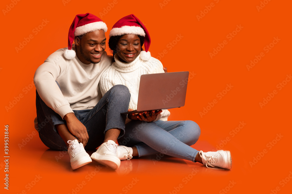 Happy Black Couple In Santa Hats Sitting With Laptop Over Orange Background Happy Black Couple In Santa Hats Sitting With Laptop Over Orange Background