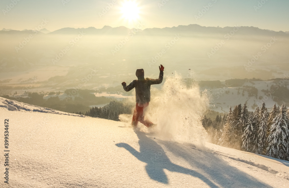 Happy Man with hood running in deep powder snow with snowshoes at sunrise. Snow is spraying and splashing. Allgau Alps, Bavaria, Germany. Happy Man with hood running in deep powder snow with snowshoes at sunrise. Snow is spraying and splashing. Allgau Alps, Bavaria, Germany.