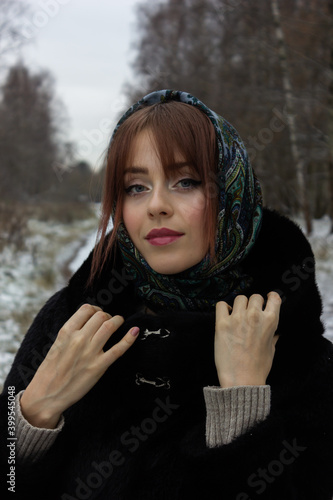 A Russian girl in a black fur coat and a green scarf in the winter looks at the camera.