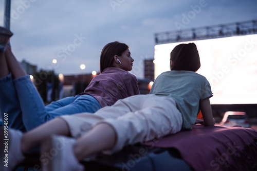 Two girls lying on the roof of a car, having fun while watching a movie in an open air cinema with a big white screen