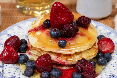 Stack of homemade pancakes prepared with blueberries, strawberries and maple syrup or honey dripping pancakes on an old plate on a wooden table. Concept healthy food at breakfast.
