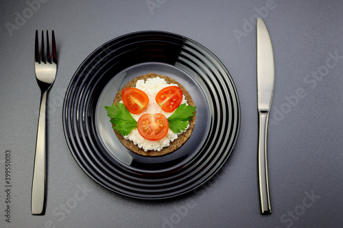 Soft fresh cheese, cherry tomatoes and celery leaves on a   slice of rye bread in a black plate on a gray background.