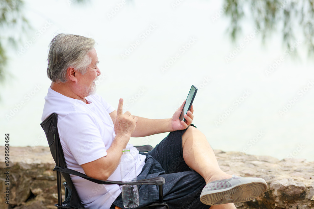 Asian senior male using mobile phone for video call communication with his family while vacation next to the beach.