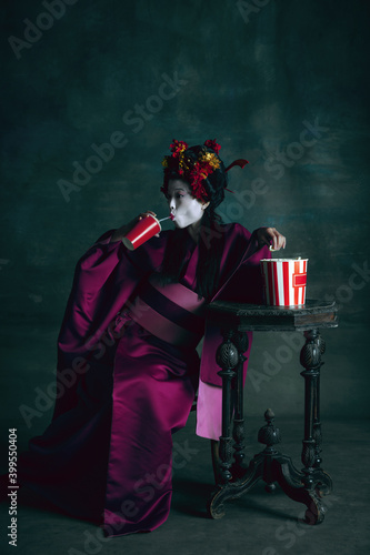 Cinema and popcorn. Young japanese woman as geisha isolated on dark green background. Retro style, comparison of eras concept. Beautiful female model like bright historical character, old-fashioned.