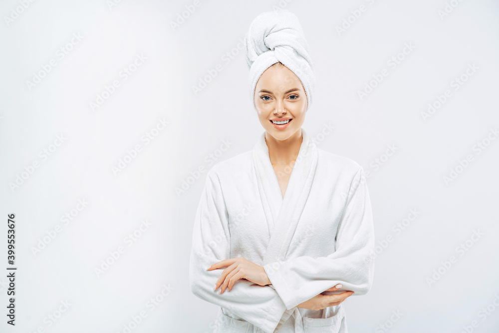 Beautiful smiling young woman has soft healthy skin after taking shower, wears bath robe and towel wrapped on head, enjoys spare time at home, isolated over white background. Wellness concept. Beautiful smiling young woman has soft healthy skin after taking shower, wears bath robe and towel wrapped on head, enjoys spare time at home, isolated over white background. Wellness concept.