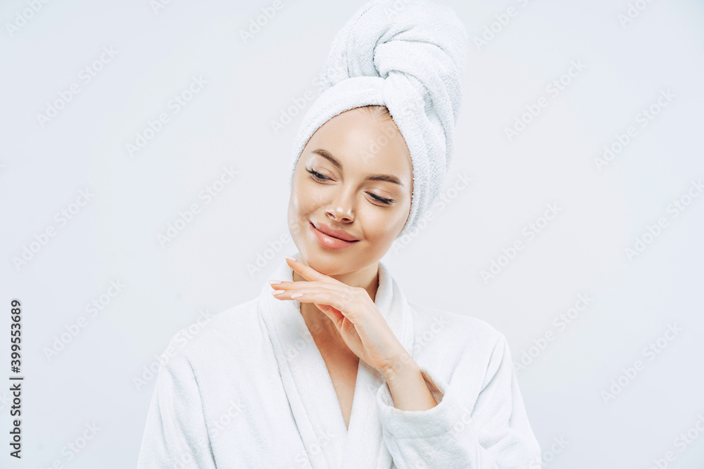 Pretty natural woman touches gently jawline, wears bath towel wrapped on head, dries hair after shower, cares about body, has clean skin, prepares for evening party, dressed in domestic robe Pretty natural woman touches gently jawline, wears bath towel wrapped on head, dries hair after shower, cares about body, has clean skin, prepares for evening party, dressed in domestic robe
