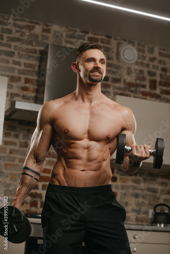 A muscular man with a beard is training his biceps with dumbbells in his apartment. A happy bodybuilder with tattoos on forearms is warming up during an arm workout at home. Athlete with a naked torso