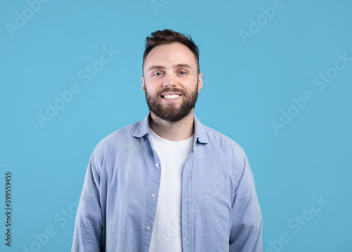 Portrait of handsome bearded young man in casual outfit smiling and looking at camera over blue studio background