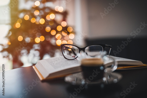 Open book, coffee and glasses on table. In the background a Christmas tree and Christmas lights. Christmas atmosphere.