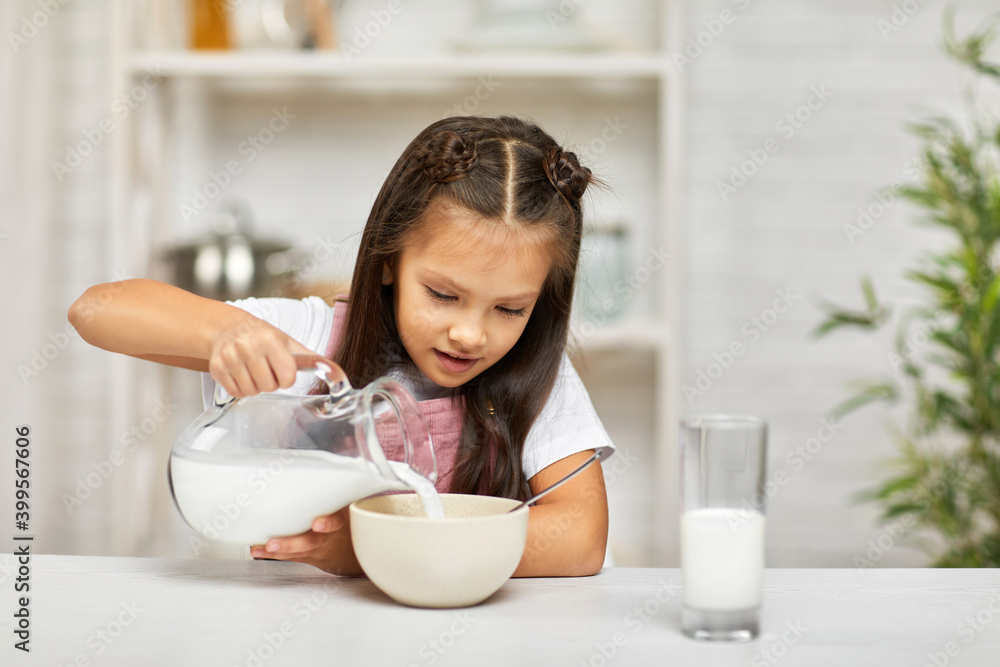 smiling cute little girl eating breakfast: cereal with the milk. child pours milk into a bowl of cereal in the kitchen. healthy breakfast