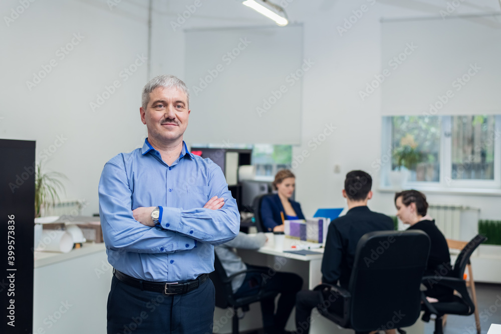 A portrait of a successful smiling senior businessman with his hands folded, his young team is on the background having a meeting in their office. A portrait of a successful smiling senior businessman with his hands folded, his young team is on the background having a meeting in their office.