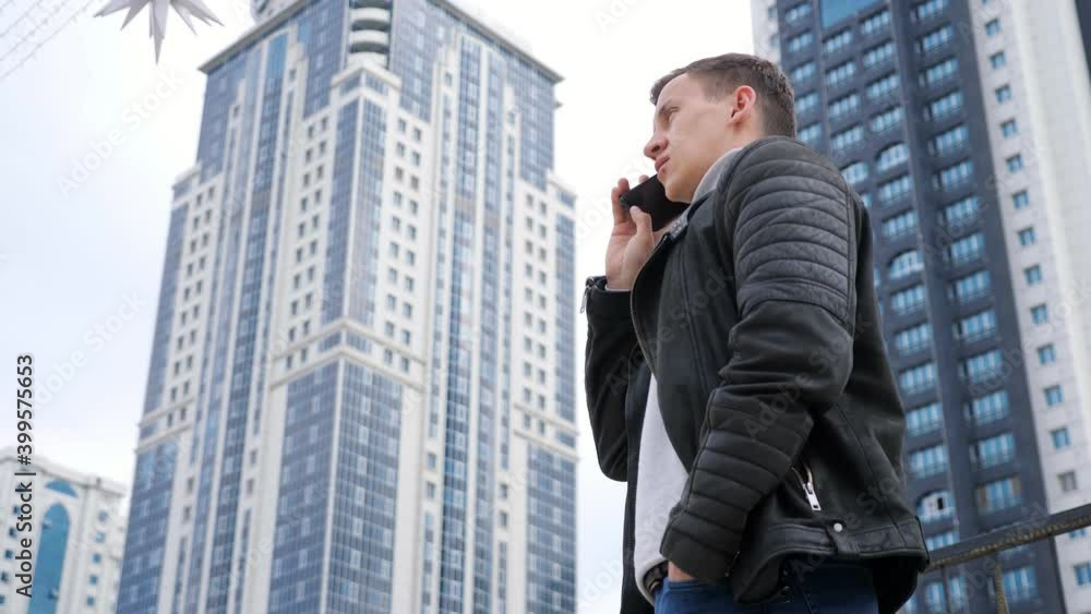 Confident businessman wearing autumn leather jacket speaks on modern smartphone standings against highrise buildings close low angle shot Confident businessman wearing autumn leather jacket speaks on modern smartphone standings against highrise buildings close low angle shot