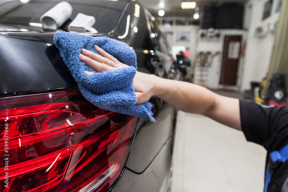 A male worker washes a black car, wiping water with a soft cloth and microfiber, cleaning the surface to shine in a vehicle detailing workshop. Auto service industry.