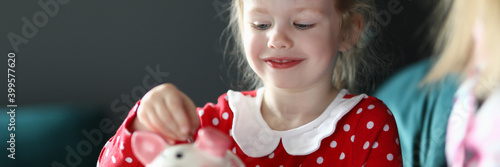 Portrait of smiling beautiful child putting coin in piggy bank sitting on sofa. Lovely kid saving up for future on toy. Girl in pretty summer dress. Childhood and happiness concept photo