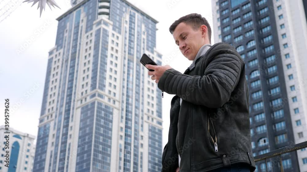 Concentrated handsome man wearing jacket surfs internet in smartphone standing against highrise buildings of residential complex low angle shot Concentrated handsome man wearing jacket surfs internet in smartphone standing against highrise buildings of residential complex low angle shot