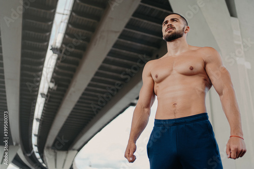 View from bottom of self confident bearded handsome European man has strong perfect body looks into distance, leads active lifestyle, poses outside under bridge. Young shirtless bodybuilder.
