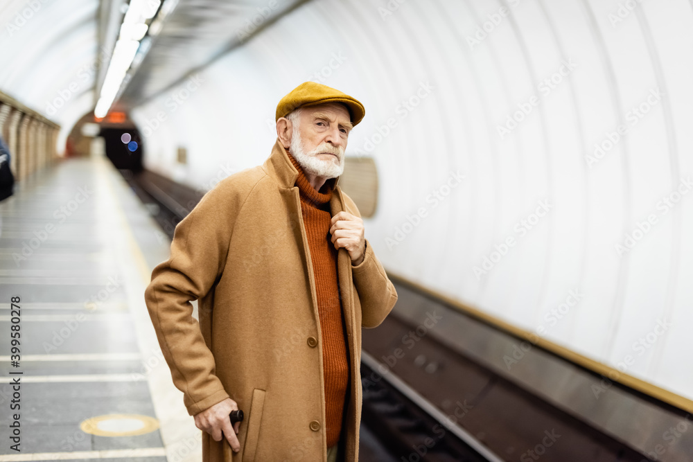 senior man in autumn outfit touching collar of coat while looking away on metro platform