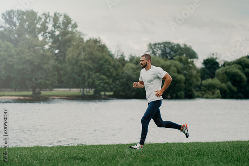 Sporty European man dressed in t shirt, trousers and sneakers and runs along river, trains outdoors, leads active lifestyle, breathes fresh air. People, sport, wellness and recreation concept.