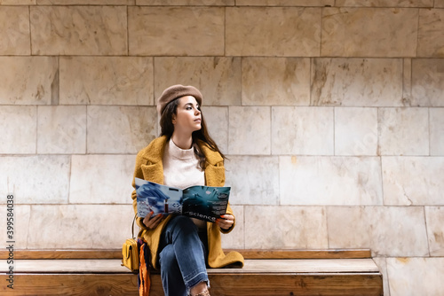stylish woman in autumn outfit looking away while holding magazine on subway platform bench