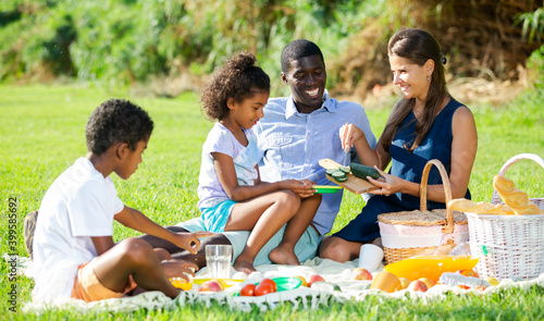 Portrait of cheerful interracial family with two children enjoying picnic on green meadow