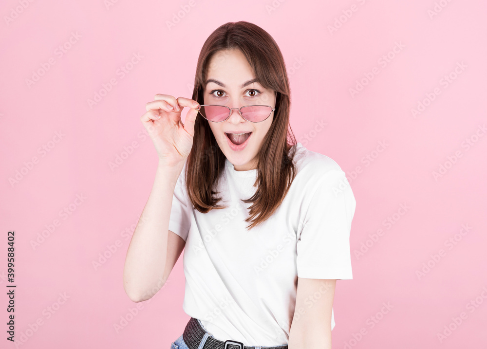 Young woman in pink glasses is very surprised looking at the camera and lowering her glasses. Surprise and shopping concept on pink background. Young woman in pink glasses is very surprised looking at the camera and lowering her glasses. Surprise and shopping concept on pink background.