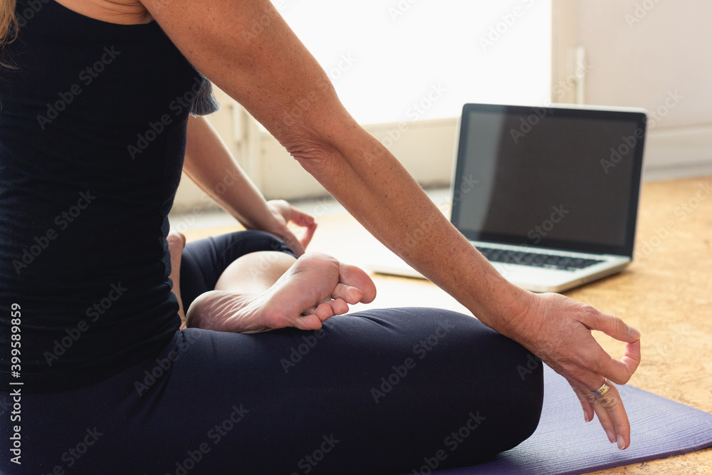 Body of slim woman sitting in padmasana in front of laptop at home. Female yogi on black outfit doing lotus pose indoors. Online yoga class, Covid-19 pandemic concepts