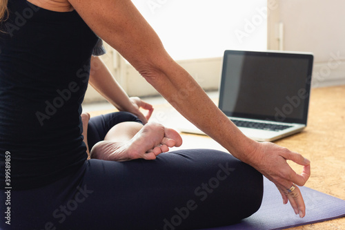 Body of slim woman sitting in padmasana in front of laptop at home. Female yogi on black outfit doing lotus pose indoors. Online yoga class, Covid-19 pandemic concepts