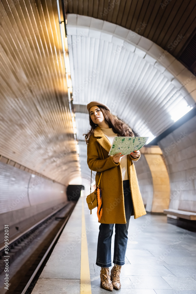 Fototapeta premium stylish woman in autumn clothes looking away while holding city map on underground platform